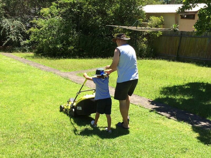 Man mowing a sunny suburban lawn with a push mower while a child helps guide it